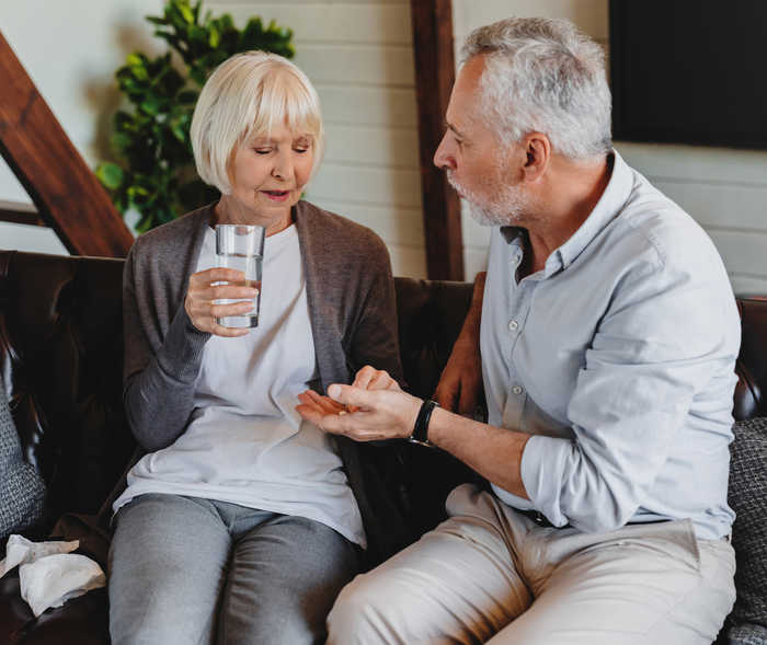 Ein älterer Mann hält eine Tablette in seiner linken Hand, die eine ältere Frau mit Wasserglas in der Hand nimmt.