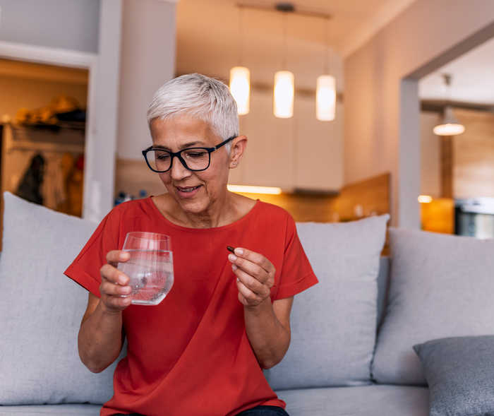 Une femme est assise sur un canapé avec un verre d’eau dans une main et une gélule dans l’autre.
