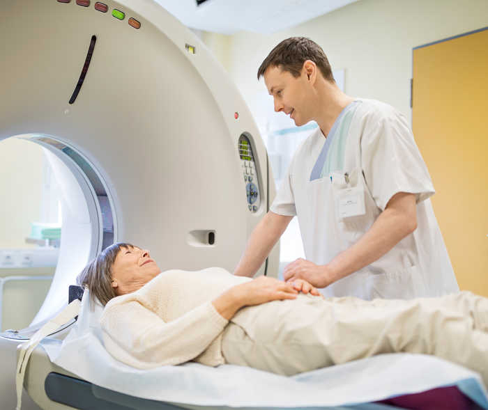Une femme âgée est sur la table d’examen pour faire un scanner CT, elle est avec un collaborateur de l’équipe de radiologie.