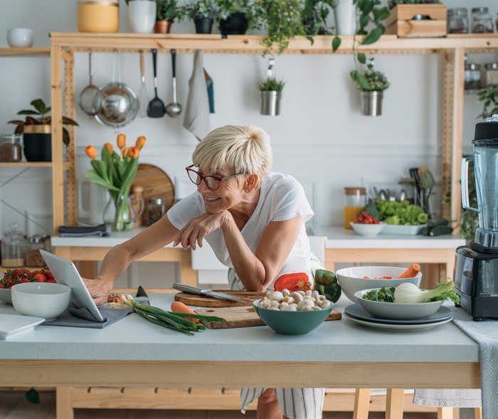 Une femme âgée prépare un plat dans la cuisine et regarde quelque chose sur une tablette.