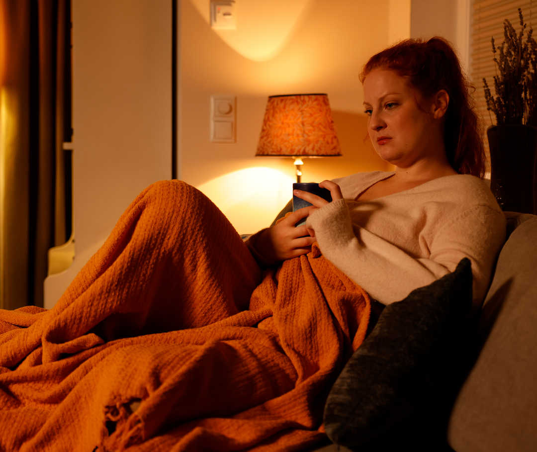 Person sitzt mit gesenktem Blick in eine Decke eingehüllt auf dem Sofa und hält eine Tasse in der Hand.