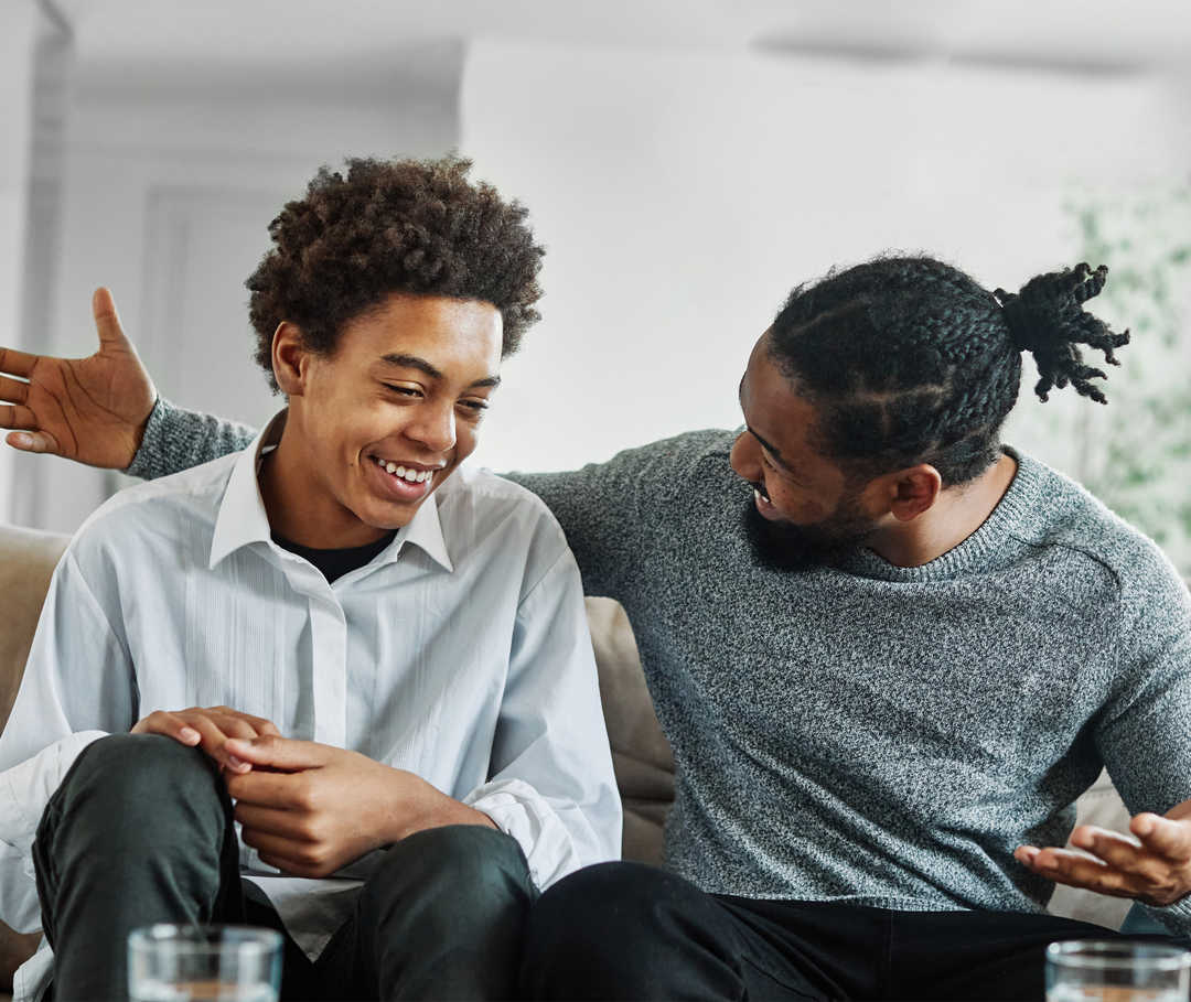 PNH in der Familie: Kindgerechte Krankheitserklärung Ein Erwachsener und ein Jugendlicher sitzen lächelnd auf einem Sofa.