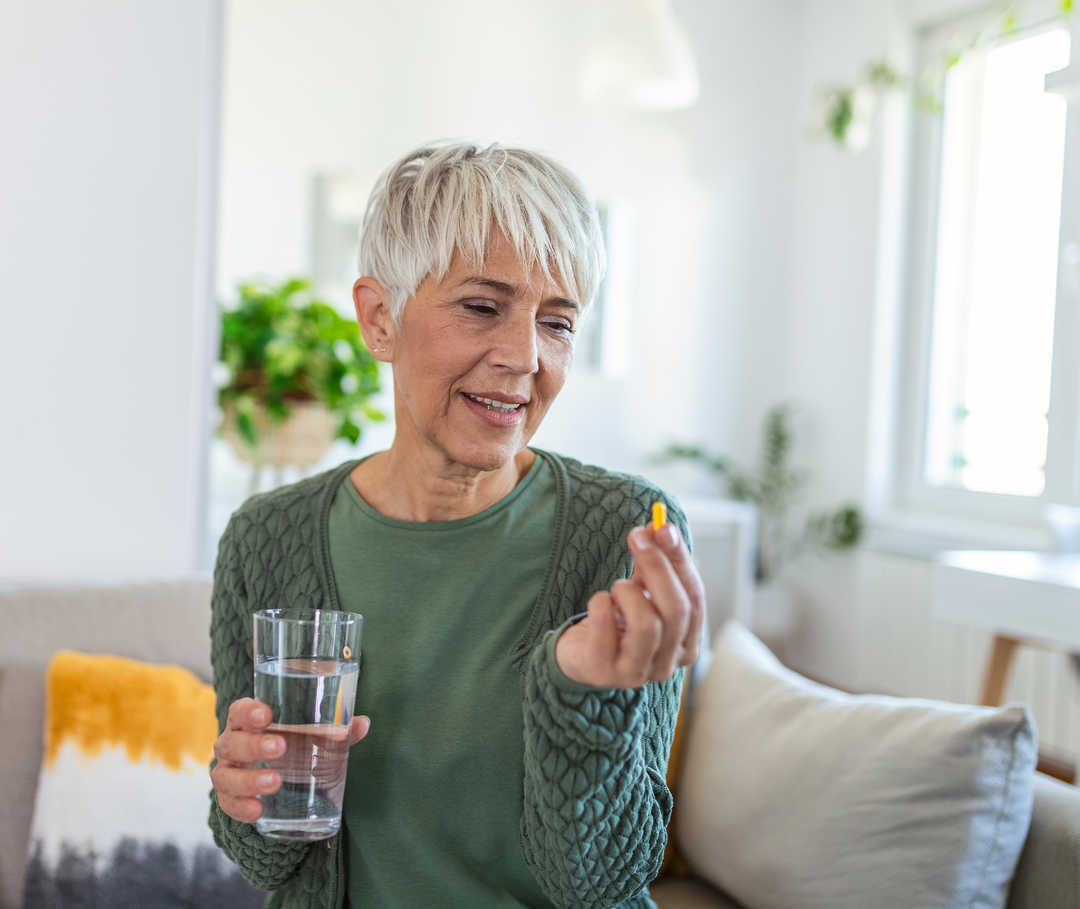 Une femme est assise sur un canapé avec un verre d’eau dans une main et une gélule dans l’autre.