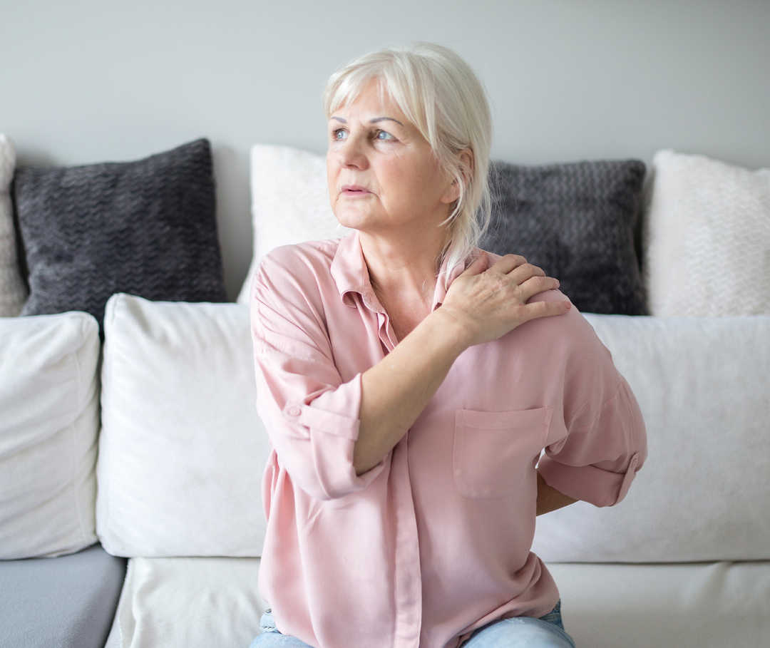 Une femme assise sur un canapé se tient l’épaule.