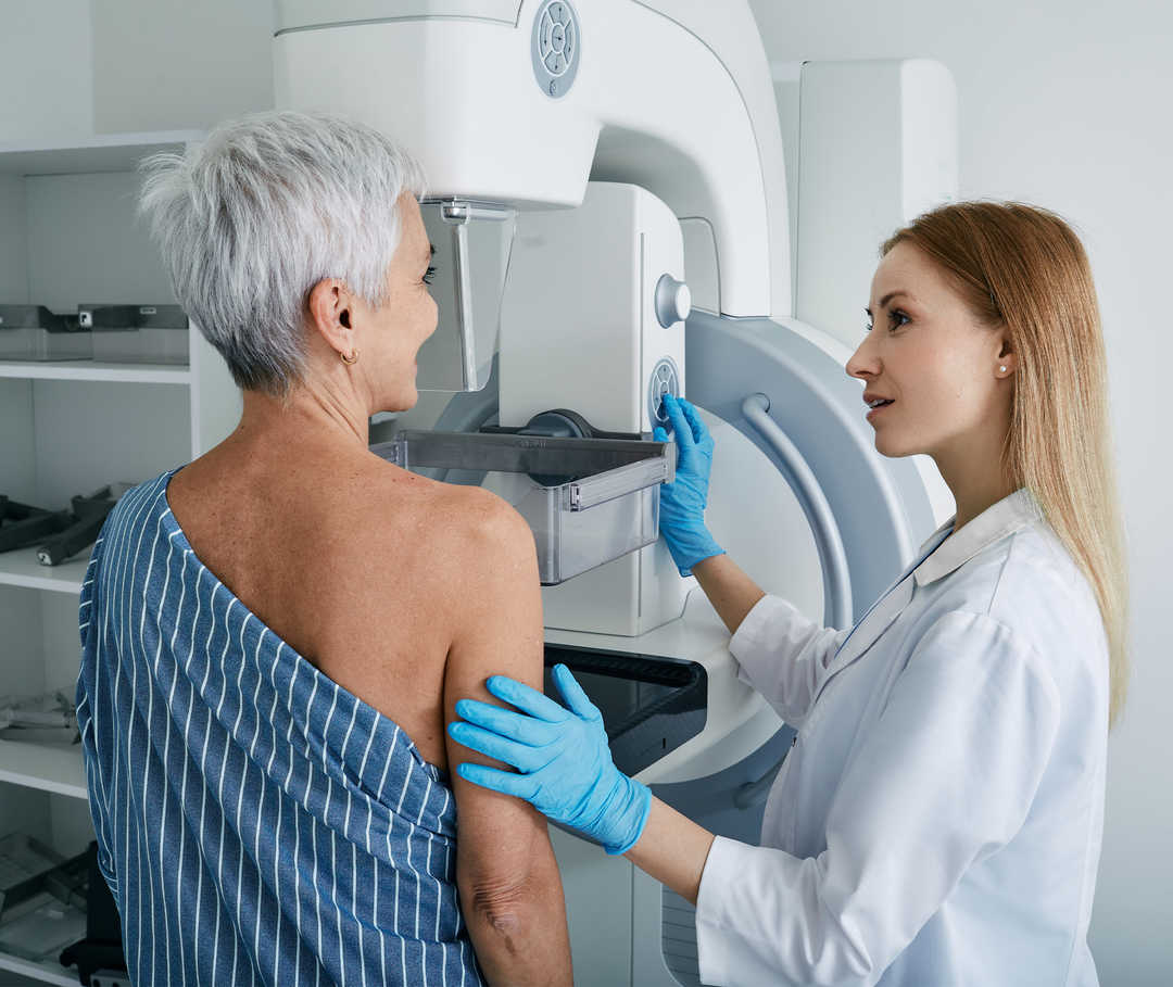 Une femme âgée se tient devant un appareil de mammographie, manipulé par une technicienne plus jeune.