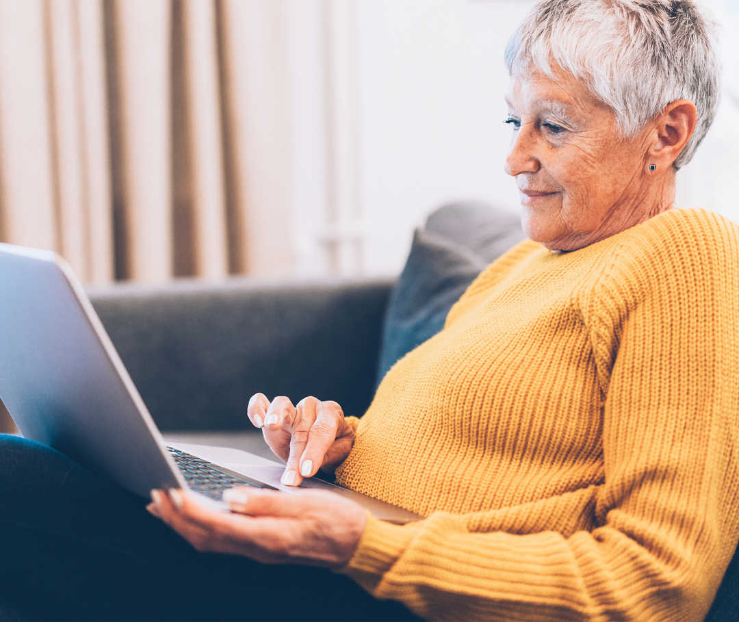 Une vieille femme est assise sur un canapé avec un ordinateur portable sur les genoux.