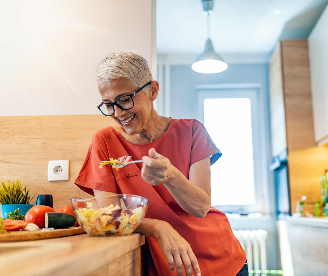 Une femme âgée est assise à la table de la cuisine, un bol de salade de pâtes devant elle, et sourit.